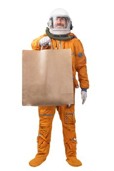 Happy astronaut wearing orange space suit and space helmet holding in hand blank kraft paper bag isolated on white background. Delivery concept. Astronaut courier concept