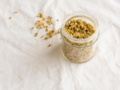 High angle view of dried chamomile flowers for tea in glass jar and scattered on white tablecloth (selective focus)