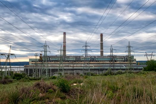 Thermal power station. Industrial landscape with thermal power plant, pipes with smoke, buildings, green trees, blue sky with clouds at sunset in summer. 