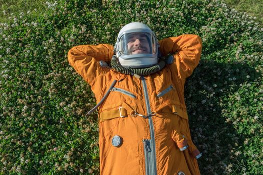 Happy cosmonaut wearing helmet with sunglasses and space suit having a rest while lying on green grass outdoor. Sunglasses concept