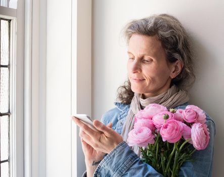 Middle aged woman with grey hair and denim shirt using smart phone, holding pink flowers and smiling