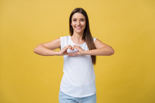 Portrait of young, brunette, pretty girl in white shirt and jeans showing with her finger heart figure, sending love to her lover, standing over yellow background.