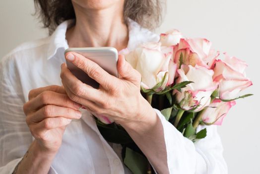 Middle aged woman in white shirt holding smart phone and pink and cream roses (cropped and selective focus)