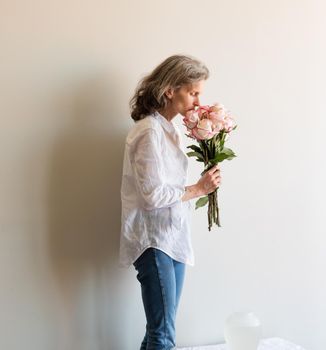 Middle aged woman with grey hair and white shirt smelling pink and cream roses near vase on table