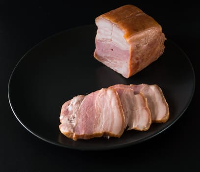 Food, a piece of bacon and cut into slices on a black plate on a black background