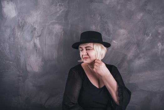 A blonde, short-haired, chubby older woman in black clothes and a black hat on a solid background of a dark gray concrete.