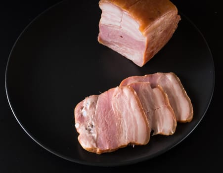 Food, a piece of bacon and cut into slices on a black plate on a black background