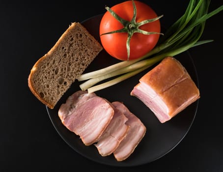 Food, Sliced bacon, tomato green onion and a piece of black bread on a black plate on a black background