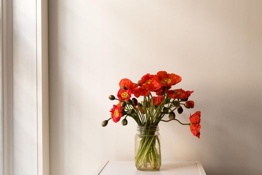 Red poppies in glass jar on small white table next to window with moody lighting