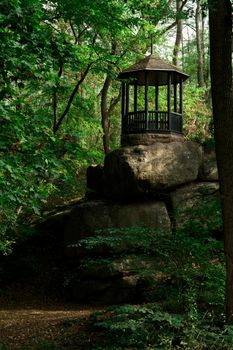 Forest park area, in the center there is a gazebo on a rock