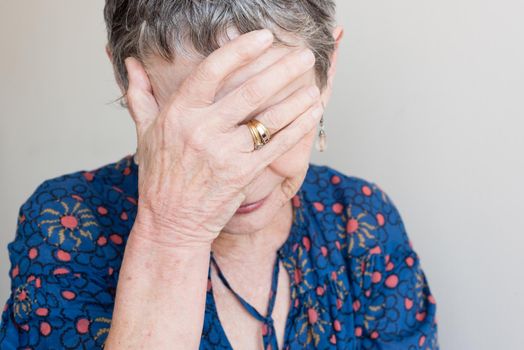 Closeup of older woman in blue top with hand covering face against  neutral background (selective focus)