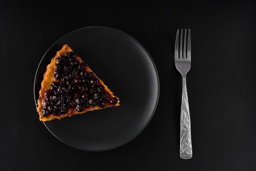 Piece of pie with carrots and currants sprinkled on top on a black plate and a cutlery fork, on a black background