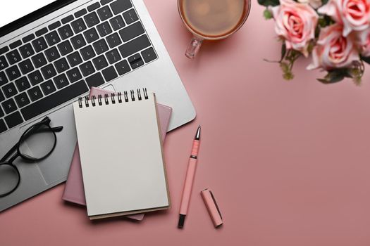 Laptop computer, coffee cup, blank notepad and flowers on pink background. Flat lay, Top view, Copy space.