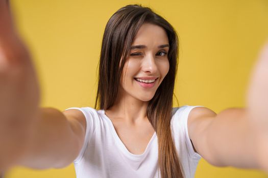 Pleasant attractive girl making selfie in studio and laughing. Good-looking young woman with brown hair taking picture of herself on bright yellow background