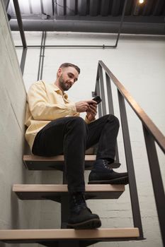 Vertical low angle shot of a man using smart phone, sitting on top of the staircase at his hotel room