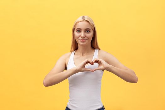 Portrait of a smiling young caucasian woman showing heart gesture with two hands and looking at camera isolated over yellow background.