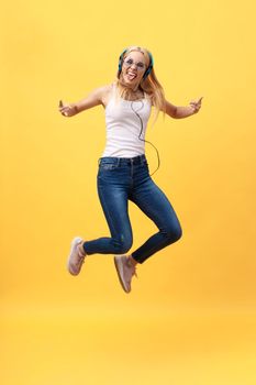 Full-length portrait of carefree woman in jean jumping while listening music. Indoor photo of adorable caucasian female model in white t-shirt fooling around in studio with yellow background