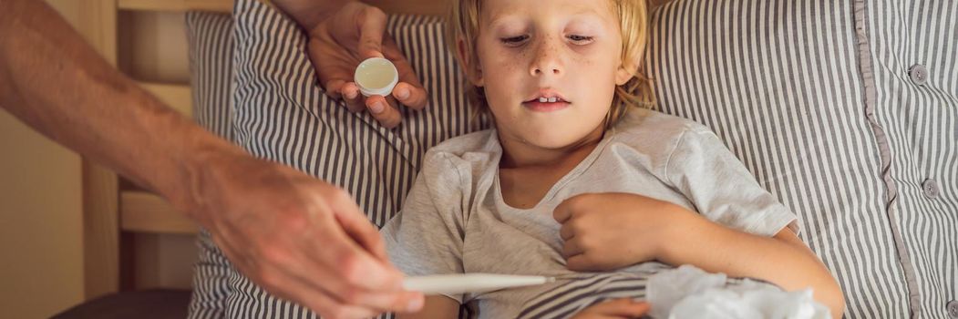 BANNER, LONG FORMAT Sick boy with thermometer laying in bed and father hand taking temperature. father checking temperature of her sick son who has thermometer. Sick child with fever and illness in bed.