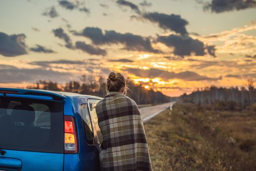 A woman in a plaid stands by the car on the side of the road in the background of the dawn. Road trip concept.