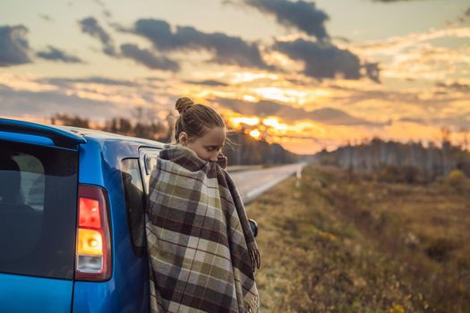 A woman in a plaid stands by the car on the side of the road in the background of the dawn. Road trip concept.