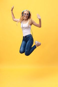 Full-length portrait of carefree woman in jean jumping while listening music. Indoor photo of adorable caucasian female model in white t-shirt fooling around in studio with yellow background