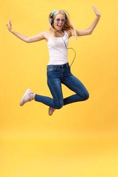 Full-length portrait of carefree woman in jean jumping while listening music. Indoor photo of adorable caucasian female model in white t-shirt fooling around in studio with yellow background