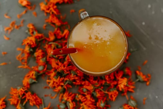 A small cup is filled to the top with yellow liquid honey. Medicinal herbal dried plants marigold, orange calendula. Neutral white and gray background.