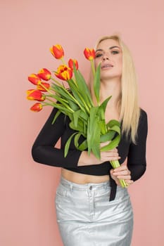 Fashion model woman in fashionable clothes on pink background. Wearing stylish clothing, black blouse, silver skirt. Posing in studio. Holding red tulips in her hands