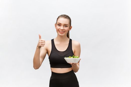 Woman in sportswear with salad, showing thumb up, isolated over white background. Young sporty blond model at studio shot. Health, beauty and dieting concept.