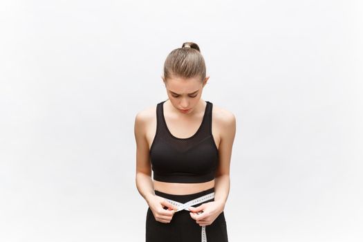 Young woman with excess weight in sporty top sadly looking on result of measuring waist over white background.