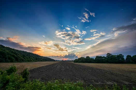 Sunset landscape of rural field in evening twilight under clouds on blue sky in bright sunset light, Colorful view of field in sunset dusk.