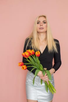 Fashion model woman in fashionable clothes on pink background. Wearing stylish clothing, black blouse, silver skirt. Posing in studio. Holding red tulips in her hands