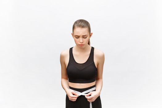 Young woman with excess weight in sporty top sadly looking on result of measuring waist over white background.