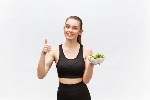 Woman in sportswear with salad, showing thumb up, isolated over white background. Young sporty blond model at studio shot. Health, beauty and dieting concept.