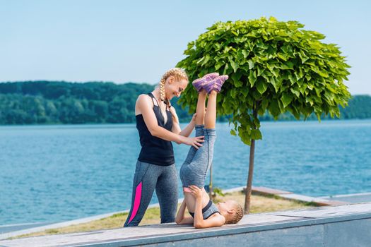 Little girt and woman are doing exercises on the grass at the shore of the lake.