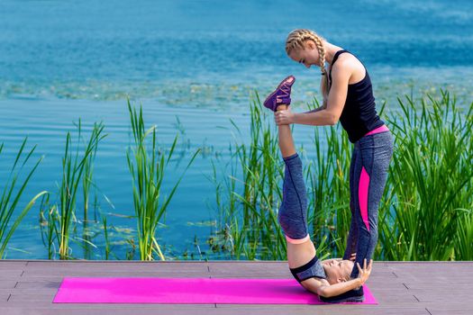 Little girt and woman are doing exercises on the grass at the shore of the lake.