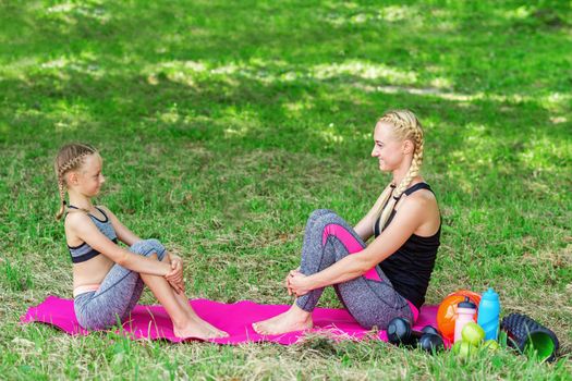Mother and her daughter are sitting on a roll mat wearing sportswear in the public park.