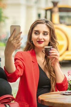 Pretty young girl is taking a selfie on a smartphone while sitting at a table at an outdoor cafe.