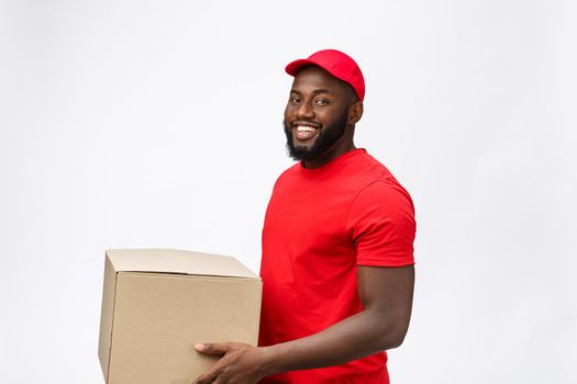 Delivery Concept - Side view Portrait of Happy African American delivery man in red cloth holding a box package. Isolated on Grey Background. Copy Space.