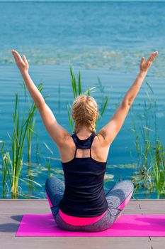 Back view of young woman is doing yoga exercise on grass of the shore of the lake.