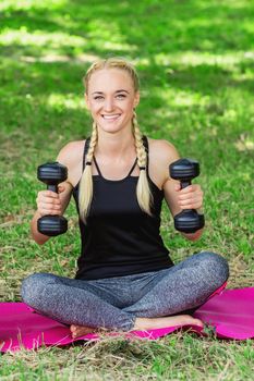 Young woman is training with black dumbbells in the park. Healthy sport exercises.