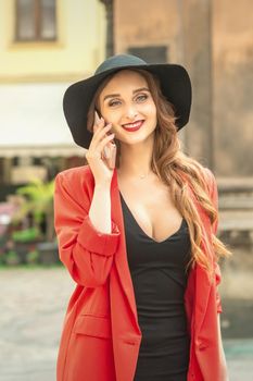 Cute fashionable young woman is talking by the phone on the street wearing hat and red jacket.