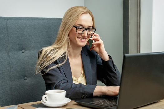 Young woman with glasses is talking by cell phone sitting at the table in home office.