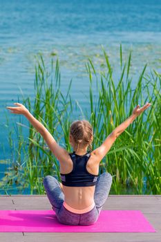 Back view of little girl is doing yoga exercise on grass of the shore of the lake.