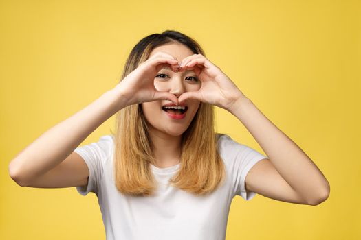 Young Asian woman show heart hand sign isolated on yellow background