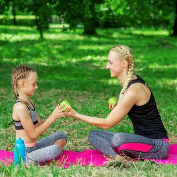 Mother and daughter snack on apples after exercise on roll mat in the park.