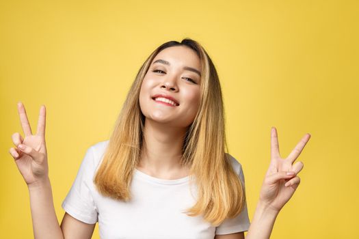 Pleased asian woman in t-shirt showing peace gestures and looking at the camera over yellow background.