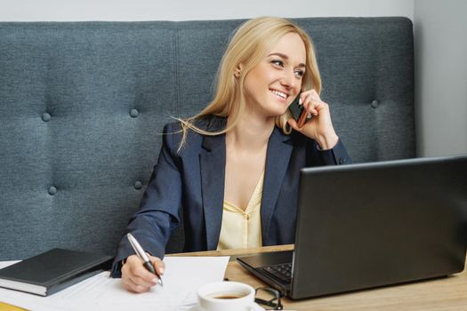 Young woman is talking by cell phone sitting at the table in home office.