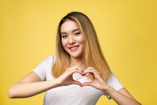 Young Asian woman show heart hand sign isolated on yellow background