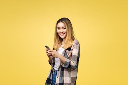 Image of satisfied asian woman with long hair smiling and texting on cell phone holding in hand isolated over yellow background.